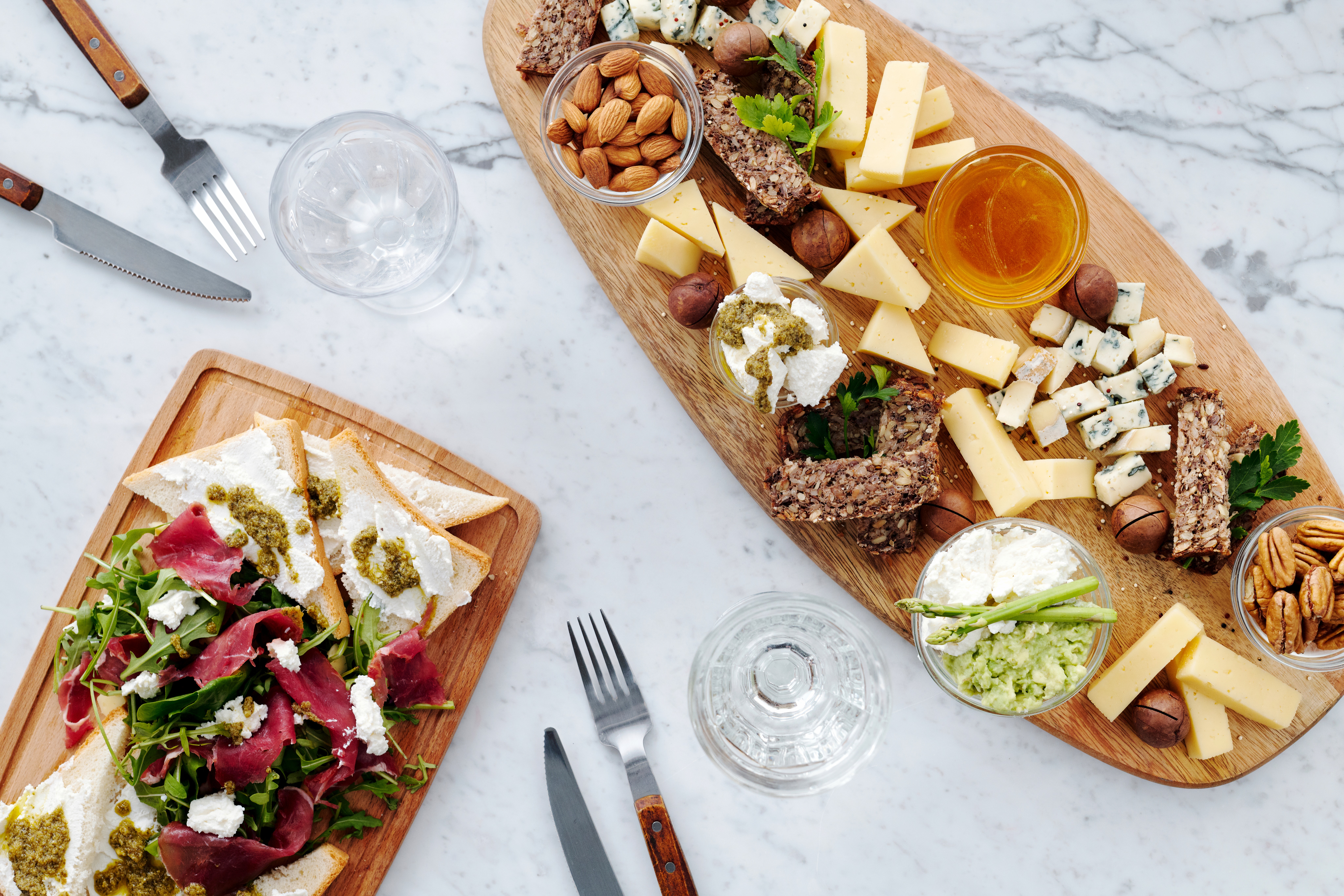 long oval charcuterie board filled with antipasto and deli items beside a rectangle wood board filled with meats, greens and breads