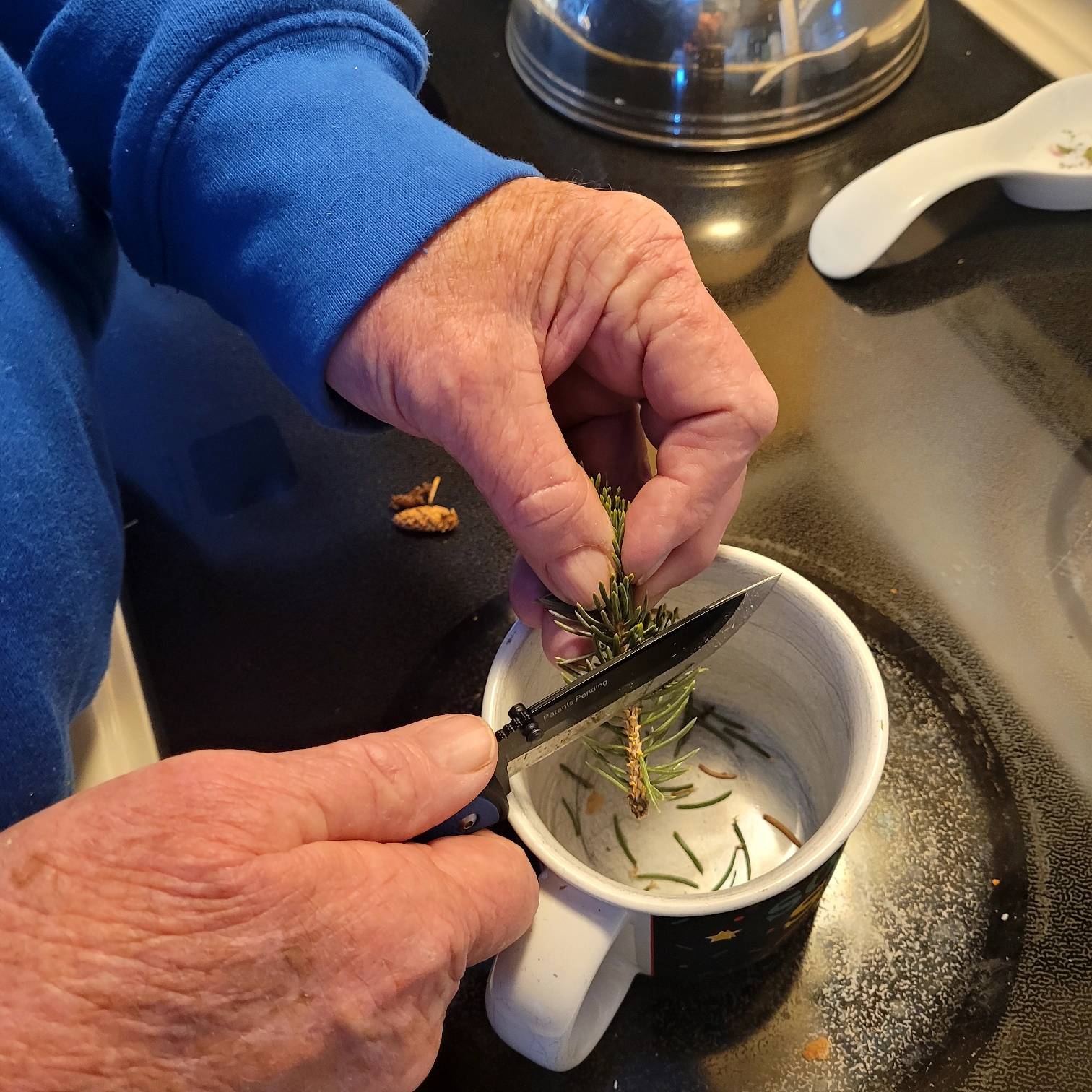 man using small knife to cut pine needles off of stem into empty ceramic mug