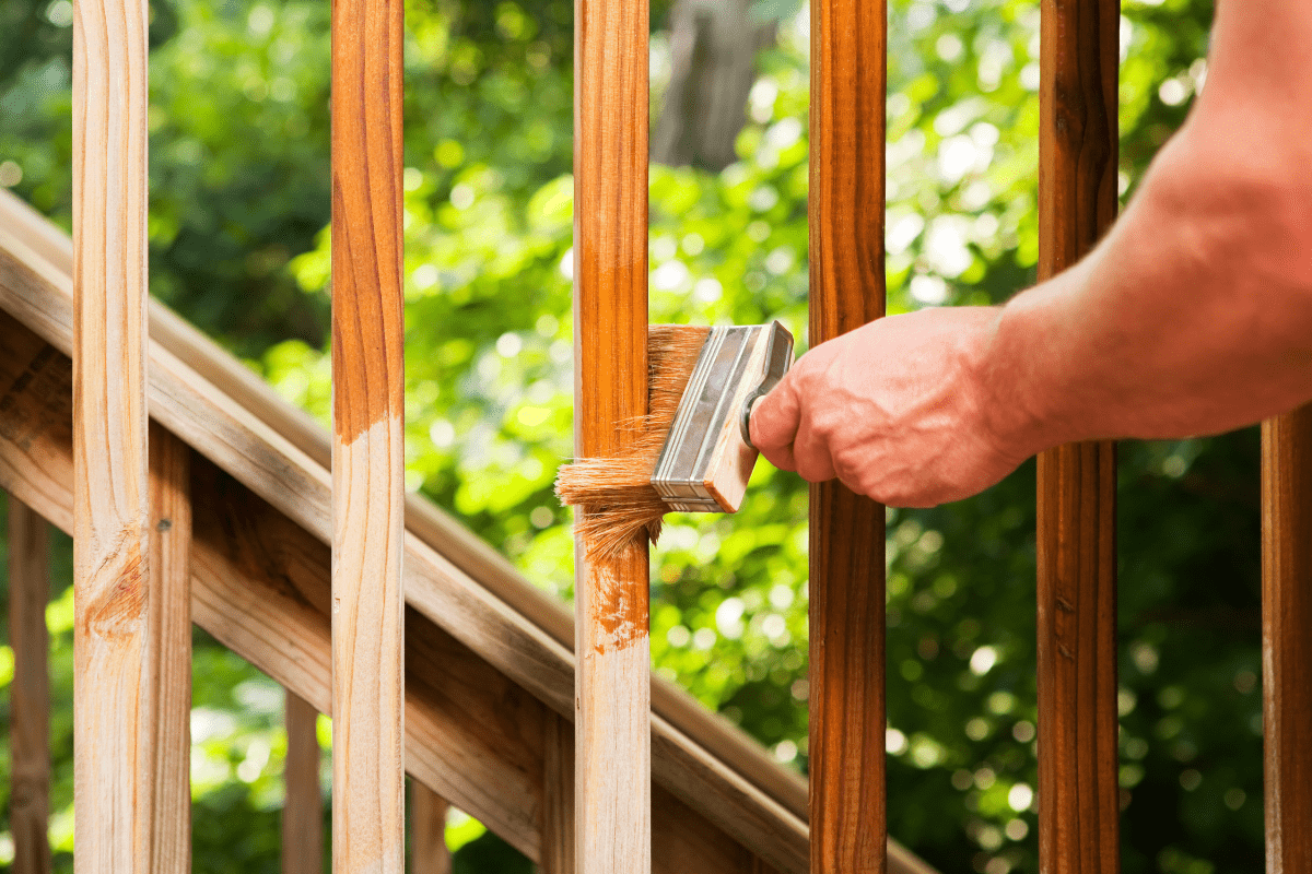 man's hand holding wide painting brush applying wood stain to spindles of deck