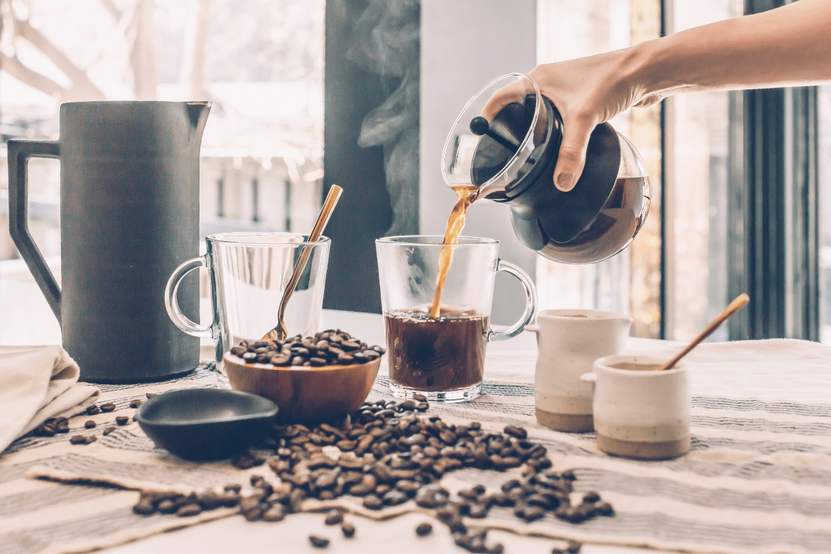 table with coffee beans spilled, ceramic mugs and jug, two glass coffee mugs with hand from out of frame pouring hot coffee