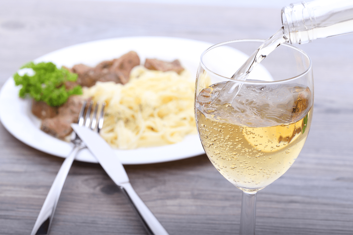 glass of white wine being poured in foreground, dinner in background