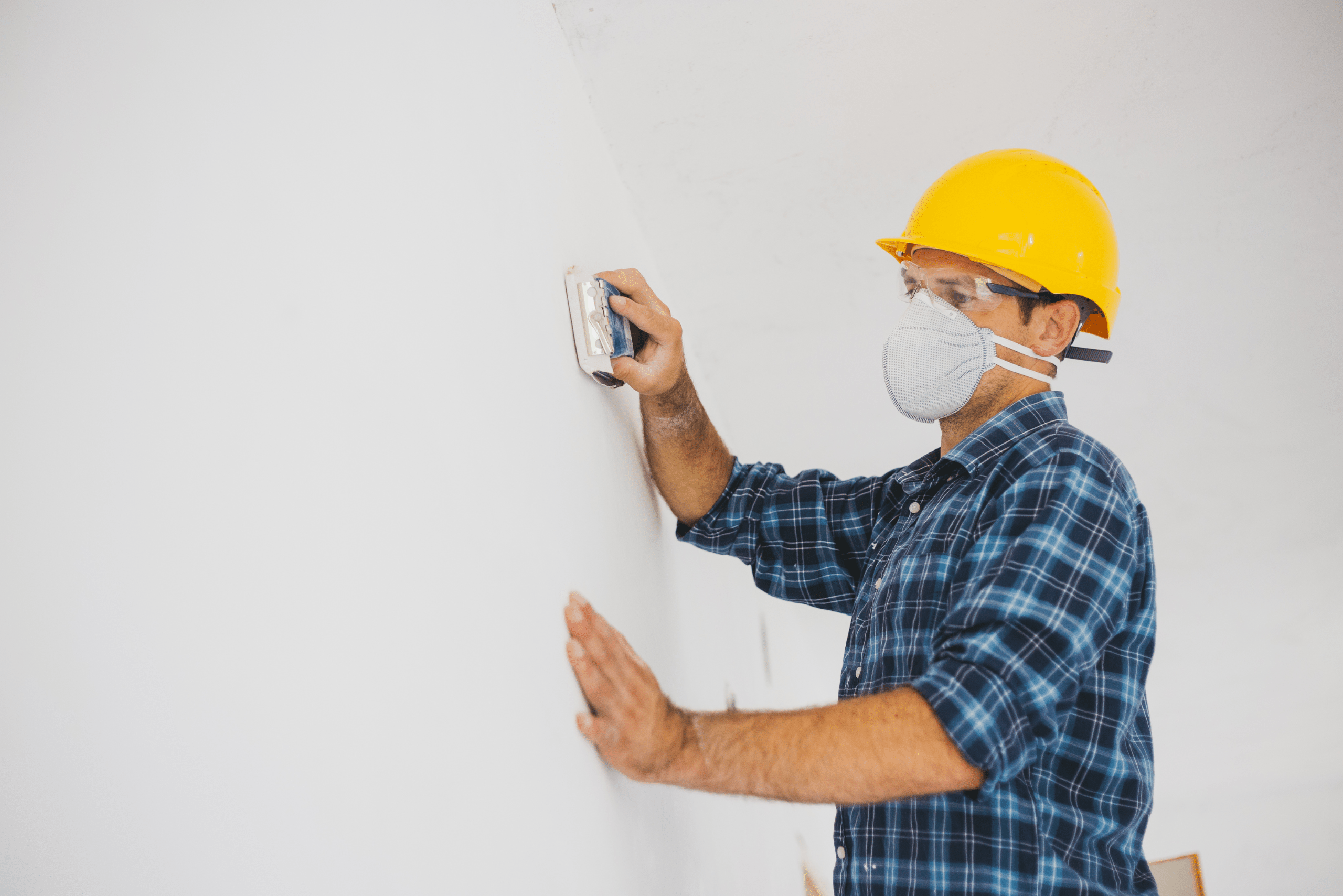 Man wearing yellow hard hat is sanding the drywall after repair.